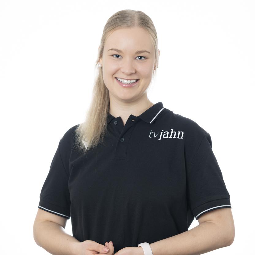 L&auml;chelnde Frau in schwarzem Poloshirt mit Logo &bdquo;tvjahn&ldquo;, steht vor hellem Hintergrund und h&auml;lt eine Hand an ihrem Oberk&ouml;rper.