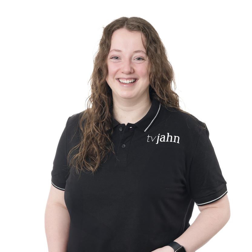 L&auml;chelnde Frau mit langen, lockigen Haaren in schwarzem Poloshirt mit Logo &bdquo;tvjahn&ldquo; vor hellem Hintergrund.