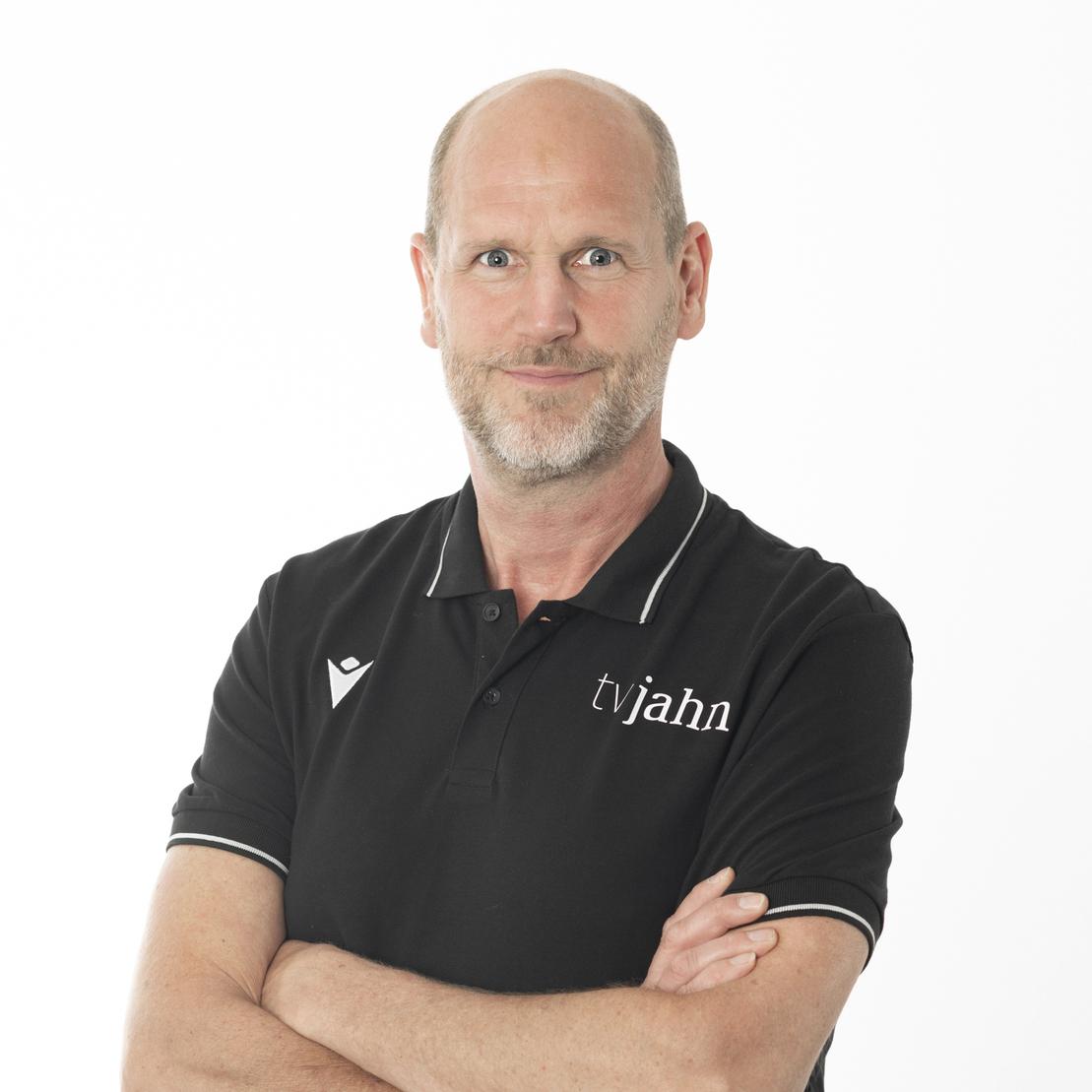 Mann in schwarzer Polo-Uniform mit "tvjahn"-Logo, l&auml;chelt und steht mit verschr&auml;nkten Armen vor hellen Hintergrund.