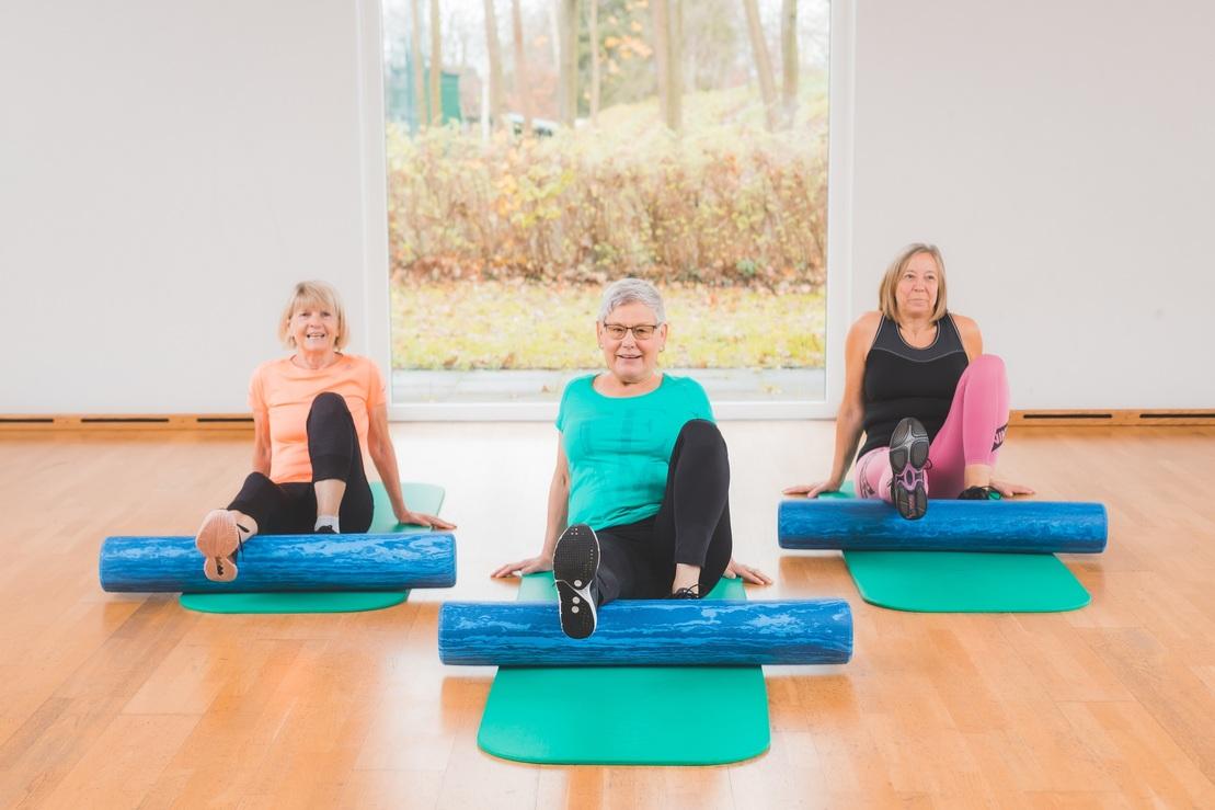 Drei Frauen sitzen auf Yogamatten und nutzen Fitnessrollen in einem hellen Raum mit Fenster.