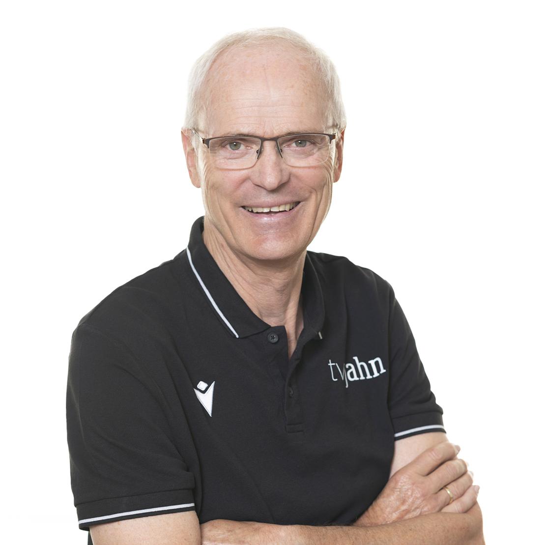 Porträt eines lächelnden Mannes mit Brille, in einem schwarzen Poloshirt mit Logo, vor schlichter weißer Hintergrund.