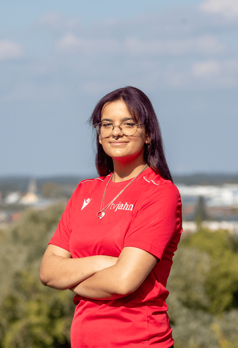 Junge Frau mit Brille und violetten Haaren, tr&auml;gt ein rotes T-Shirt, steht mit verschr&auml;nkten Armen vor einem verschwommenen Hintergrund.