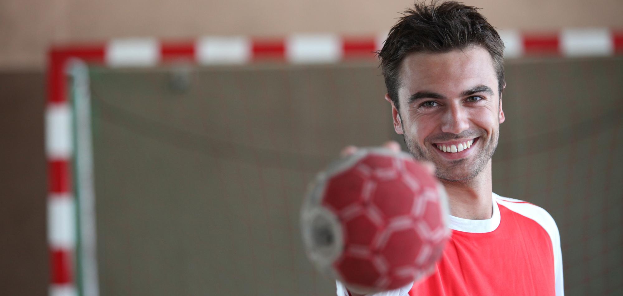 Lächelnder Handballspieler hält einen roten Handball vor sich, im Hintergrund ein Handballtor.