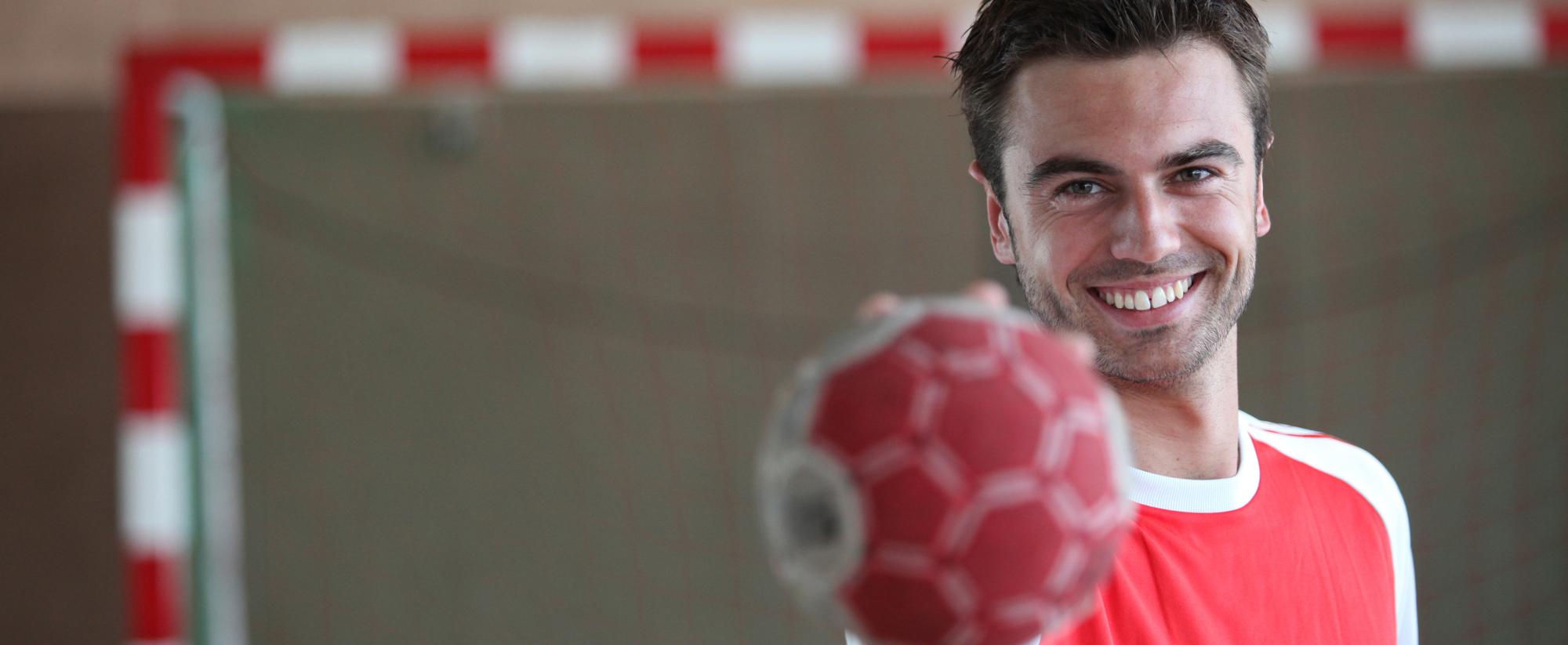 Lächelnder Handballspieler hält einen roten Handball vor sich, im Hintergrund ein Handballtor.