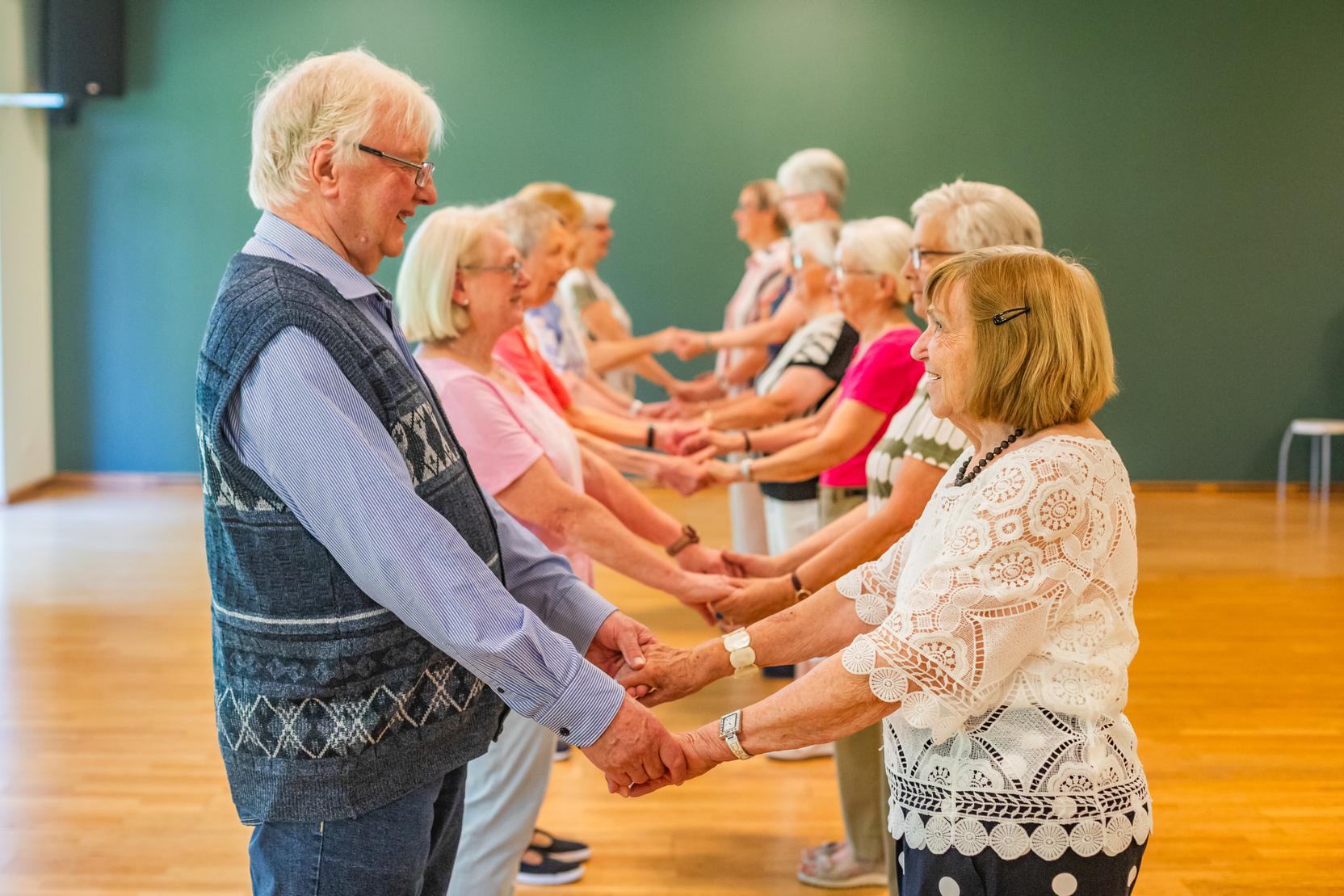 &Auml;ltere Menschen stehen in einer Reihe und halten sich an den H&auml;nden in einem lichtdurchfluteten Raum.