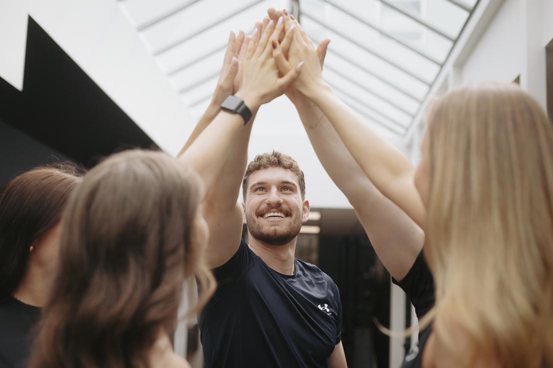 Junger Mann l&auml;chelt, w&auml;hrend er mit mehreren H&auml;nden ein gemeinsames &bdquo;High Five&ldquo; in einem hellen Raum gibt.