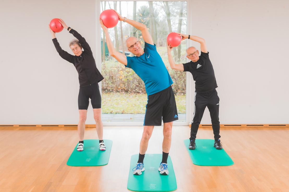 Drei Personen f&uuml;hren Dehn&uuml;bungen mit roten B&auml;llen auf gr&uuml;nen Yogamatten in einem hellen Raum aus.