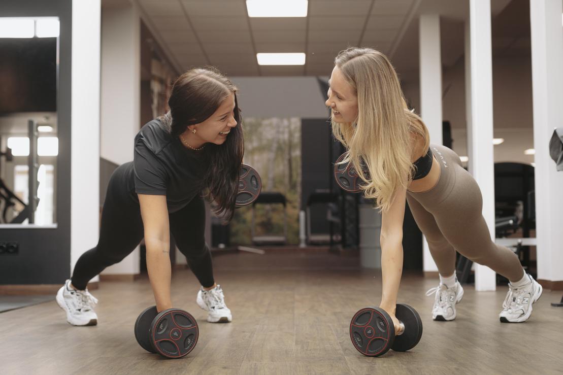 Zwei Frauen machen Plank-Übungen mit Hanteln in einem modernen Fitnessstudio.