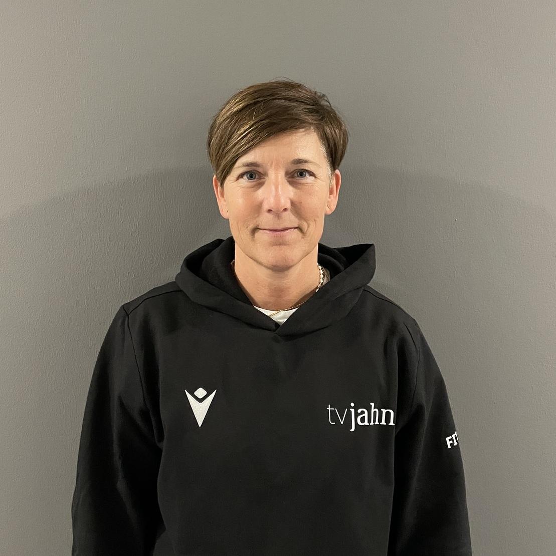 Frau mit kurzem Haar tr&auml;gt einen schwarzen Hoodie mit &bdquo;tvjahn&ldquo; und steht vor einer grauen Wand.