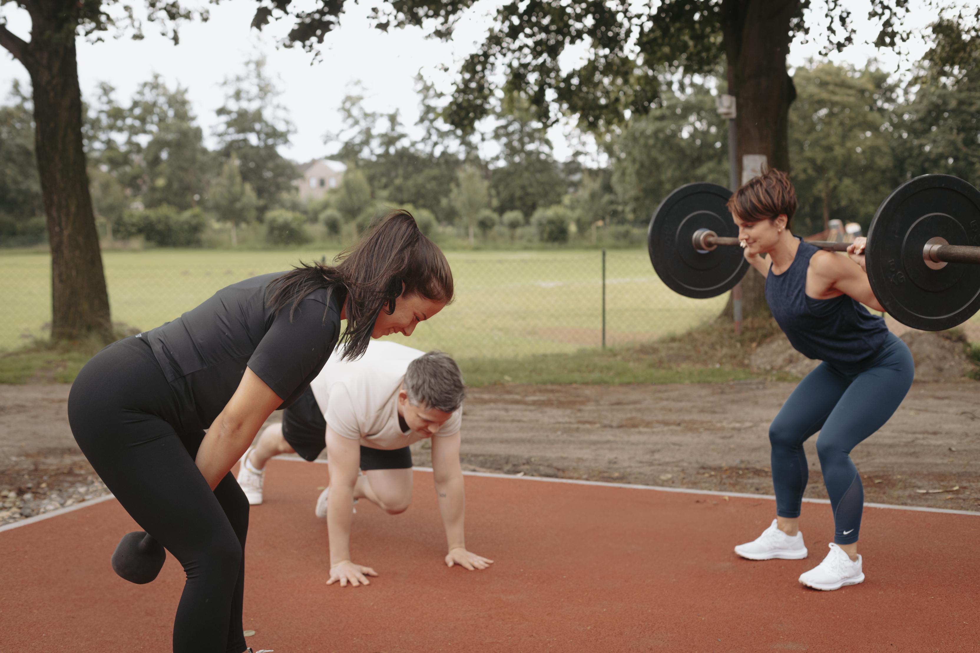 Drei Personen trainieren auf einem Außenplatz: zwei heben Gewichte, während eine Person auf dem Boden Übungen macht.