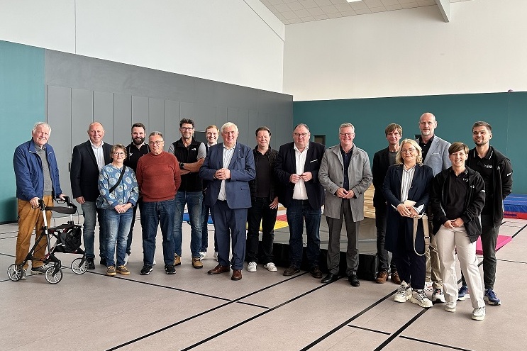 Gruppe von 15 Personen posiert in einem Sportraum mit Bodenmarkierungen und einer gr&uuml;nen Wand im Hintergrund.