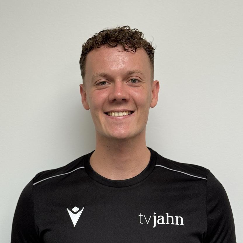 Lächelnder junger Mann in schwarzem Shirt mit "tvjahn"-Logo vor neutralem Hintergrund.