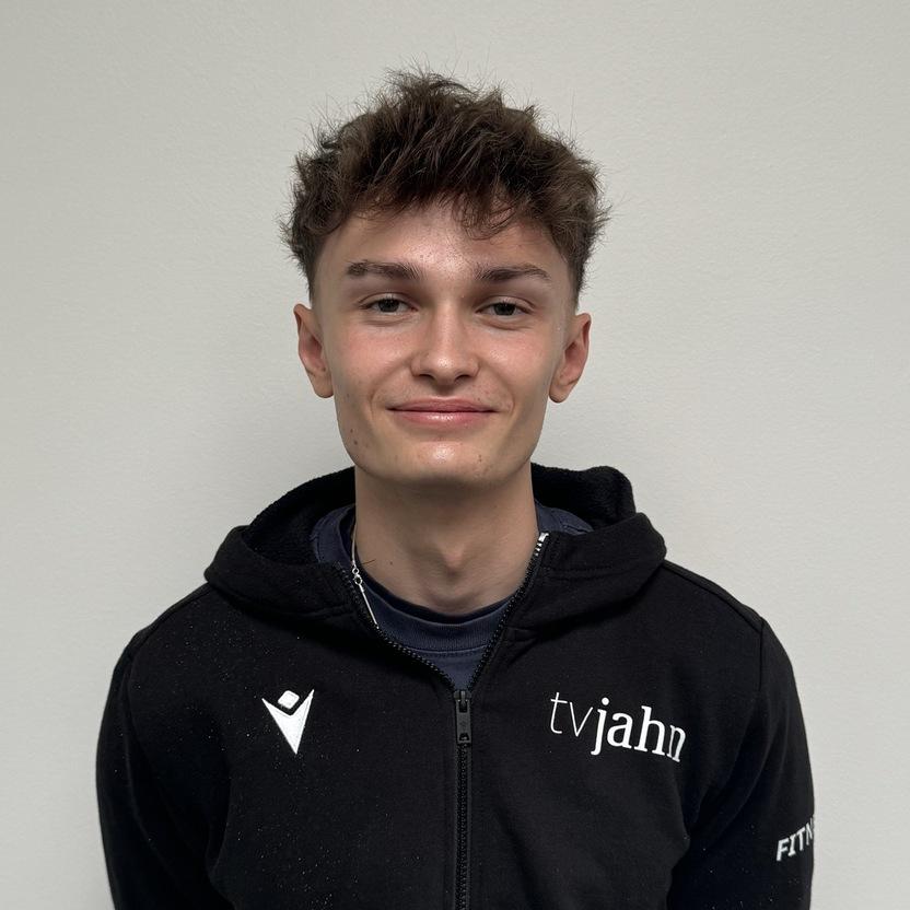 Junger Mann in schwarzer Trainingsjacke mit Logo steht vor einer hellen Wand und lächelt.