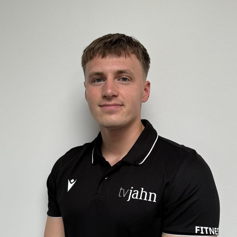 Mann in schwarzem Poloshirt mit Logo steht vor einer neutralen Wand und lächelt, die Arme vor dem Körper verschränkt.