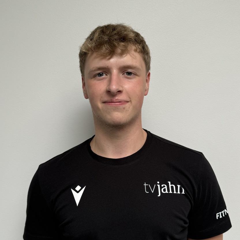 Junger Mann in schwarzem Shirt mit Aufdruck steht vor einer weißen Wand und lächelt leicht.