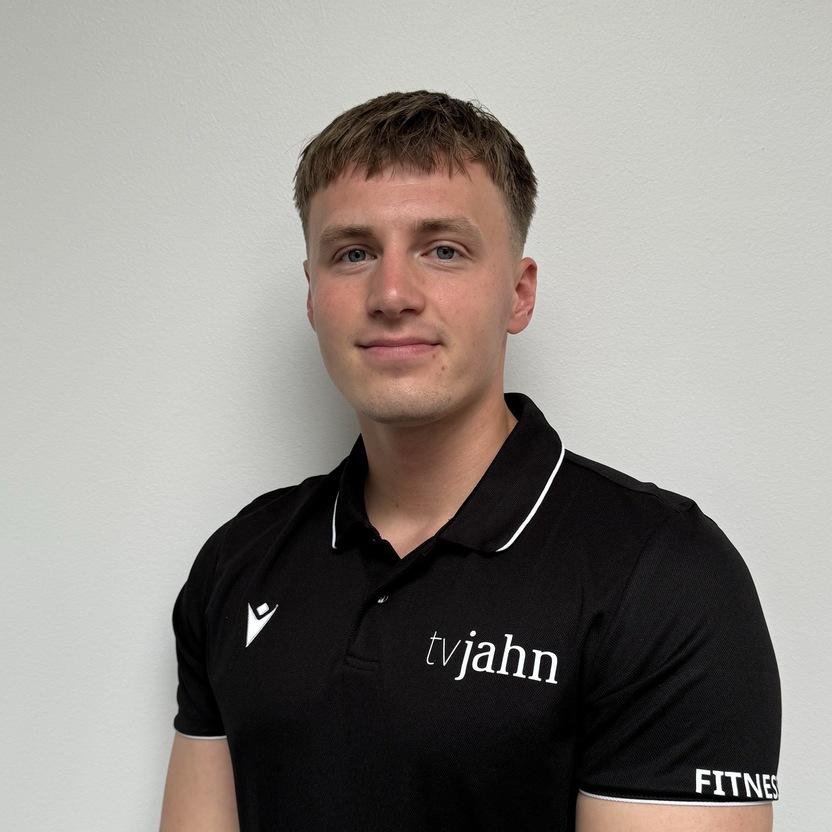 Mann in schwarzem Poloshirt mit Logo steht vor einer neutralen Wand und lächelt, die Arme vor dem Körper verschränkt.
