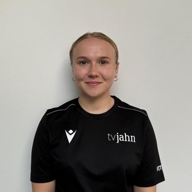 Junge Frau in schwarzem Shirt mit Logo, vor einer hellen Wand stehend, lächelt in die Kamera.