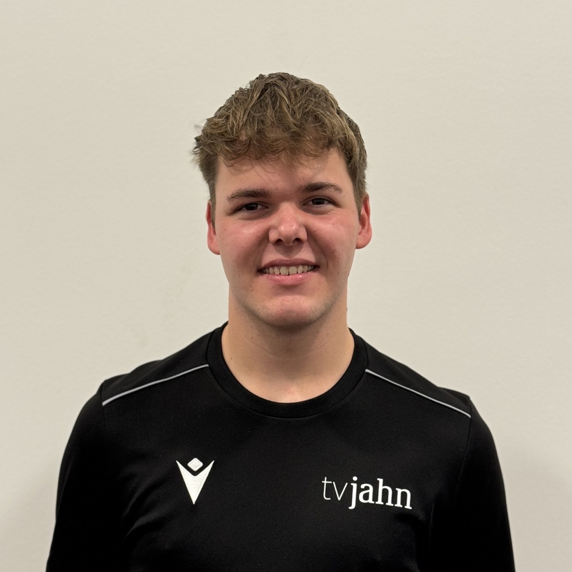 Portr&auml;t eines jungen Mannes mit kurzem Haar in einem schwarzen Shirt mit dem Aufdruck &bdquo;tvjahn&ldquo; vor einer hellen Wand.