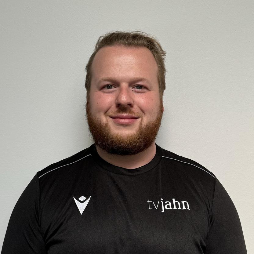 Mann mit kurzer Frisur und Bart trägt ein schwarzes T-Shirt mit dem Aufdruck "tvjahn" vor einer hellen Wand.