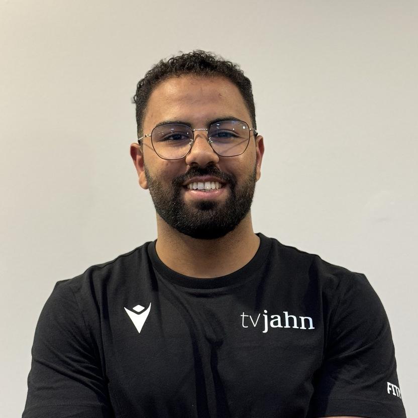Porträt eines Mannes mit Brille, der in einem schwarzen T-Shirt mit Aufschrift vor einer neutralen Wand steht.