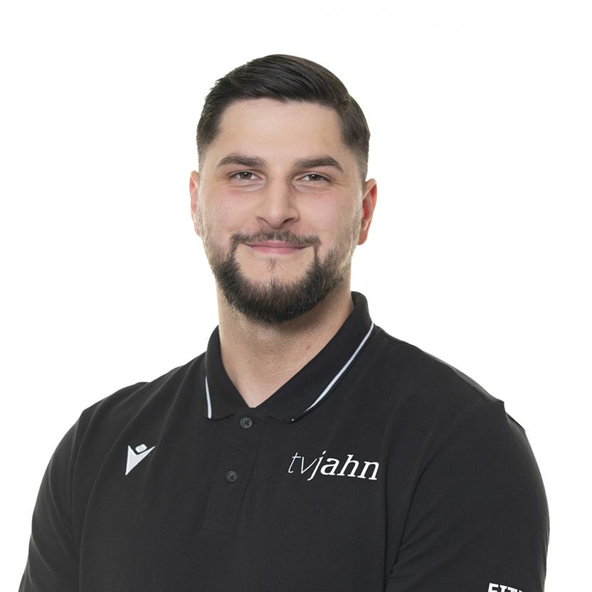 Mann mit Bart trägt ein schwarzes Polo-Shirt mit dem Aufdruck „tvjahn“ vor einem hellen Hintergrund.