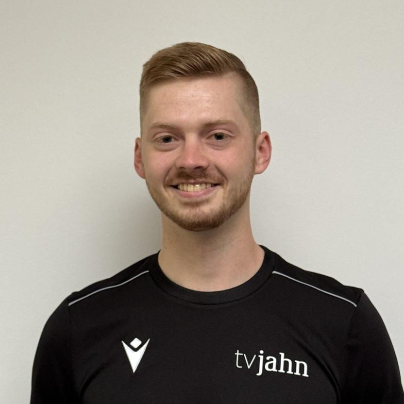 Junger Mann mit kurzem Haar in schwarzem Shirt mit Logo steht vor neutralem Hintergrund und lächelt.