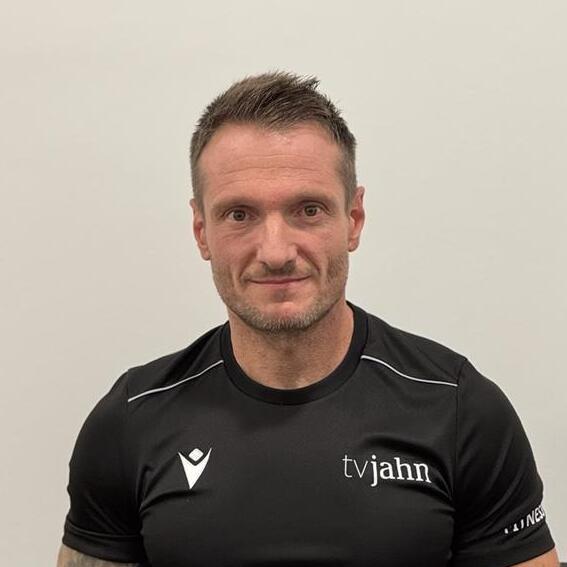 Männlicher Sportler mit kurzem Haar, trägt ein schwarzes T-Shirt mit Logo, sitzt vor einer neutralen Hintergrundwand.