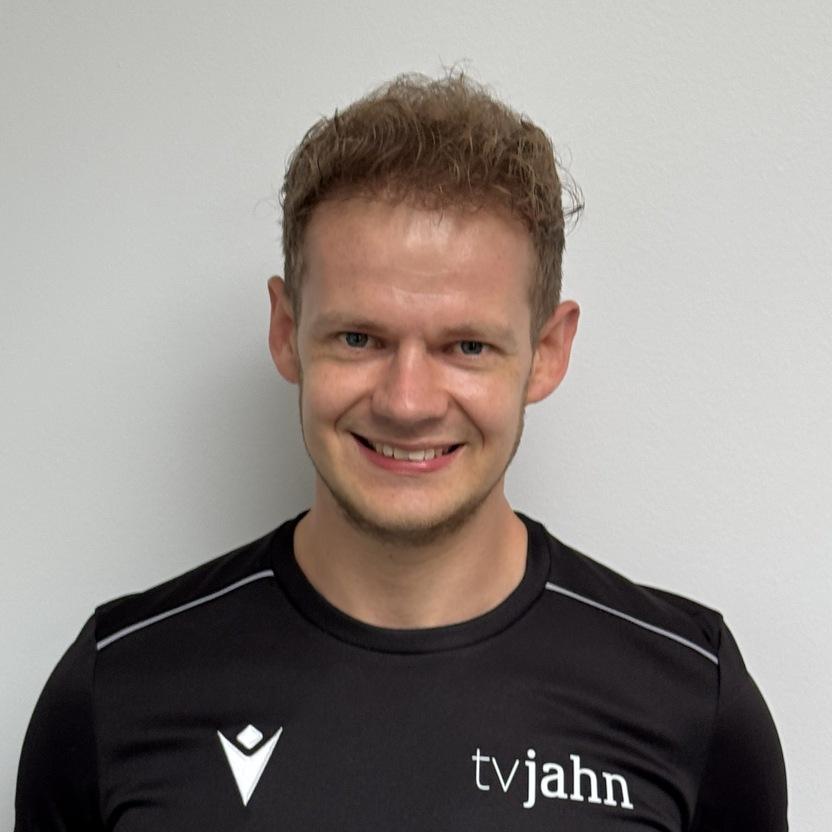 Mann mit blondem Haar trägt ein schwarzes Sportshirt mit Logo vor einer neutralen Wand. Er lächelt in die Kamera.