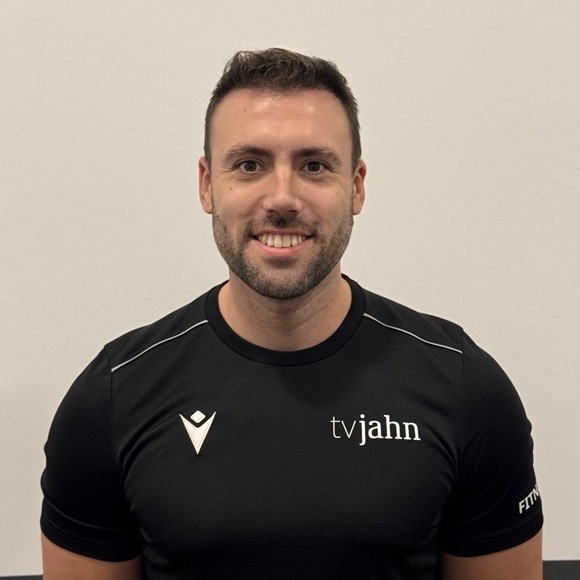 Mann mit kurzem Haar und Bart tr&auml;gt ein schwarzes T-Shirt mit dem Aufdruck &bdquo;tvjahn&ldquo; vor einer hellen Wand.