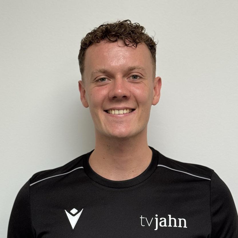 Lächelnder junger Mann in schwarzem Shirt mit "tvjahn"-Logo vor neutralem Hintergrund.