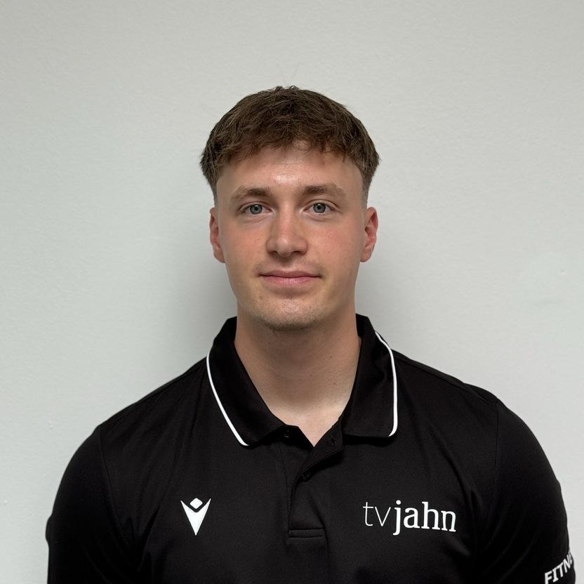 Junger Mann in schwarzem Polohemd mit Logo steht vor einer hellen Wand und blickt direkt in die Kamera.