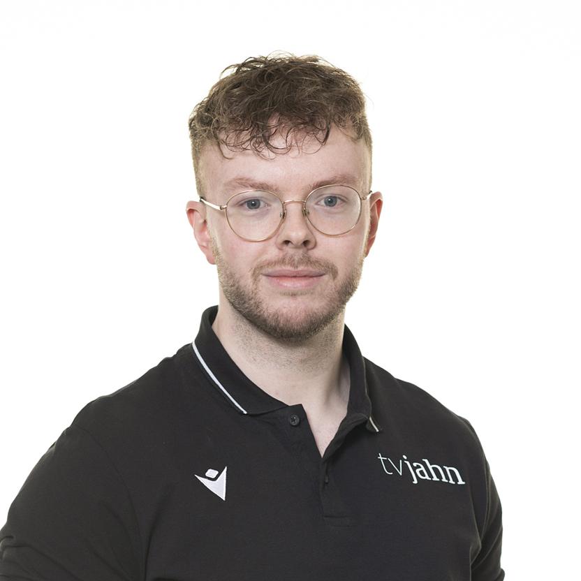 Porträt eines Mannes mit Brille, kurzem Haar und schwarzem Poloshirt mit Logo vor weißem Hintergrund.