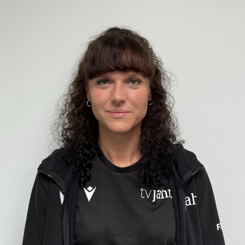 Porträt einer Frau mit lockigem, schwarzem Haar, die ein schwarzes Sportshirt trägt. Hintergrund ist eine helle Wand.