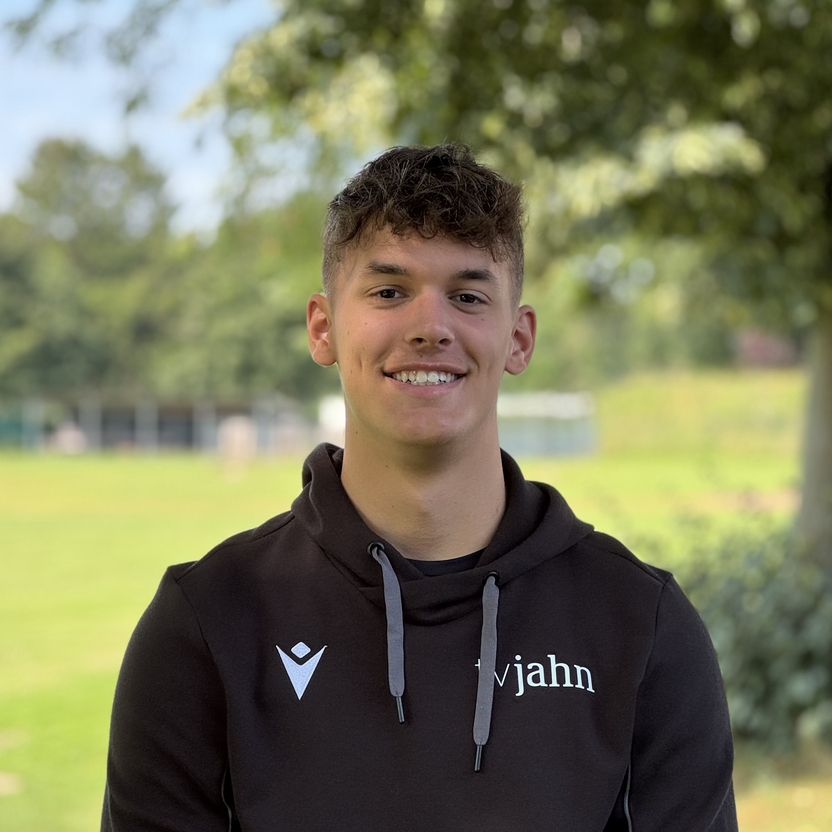 Junger Mann mit dunklem Hoodie sitzt l&auml;chelnd vor gr&uuml;nem Hintergrund mit B&auml;umen und Sportplatz.