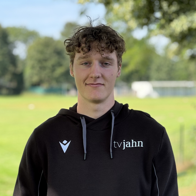 Junger Mann mit lockigem Haar in schwarzer Jacke steht vor einem gr&uuml;nen Sportplatz unter B&auml;umen.