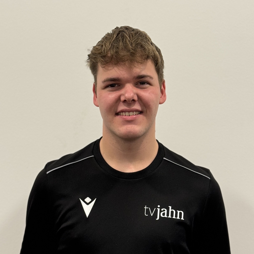 Portr&auml;t eines jungen Mannes mit kurzem Haar in einem schwarzen Shirt mit dem Aufdruck &bdquo;tvjahn&ldquo; vor einer hellen Wand.