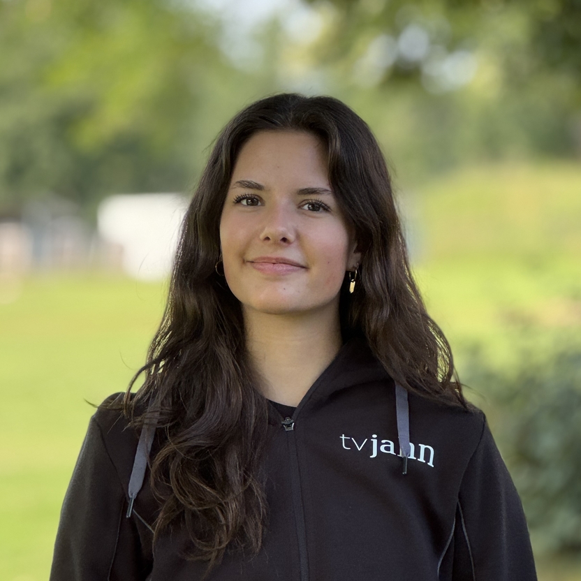 Junge Frau mit langen, lockigen Haaren tr&auml;gt einen schwarzen Hoodie mit der Aufschrift "tv jain" vor einer gr&uuml;nen Wiese.