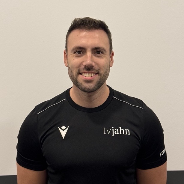 Mann mit kurzem Haar und Bart tr&auml;gt ein schwarzes T-Shirt mit dem Aufdruck &bdquo;tvjahn&ldquo; vor einer hellen Wand.