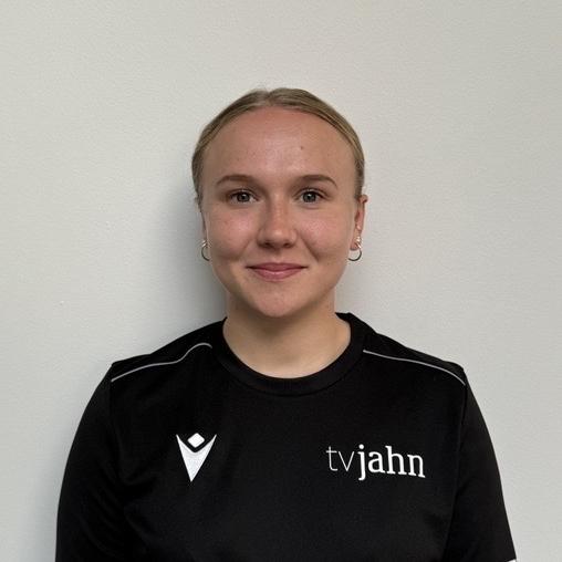 Junge Frau in schwarzem Shirt mit Logo, vor einer hellen Wand stehend, lächelt in die Kamera.