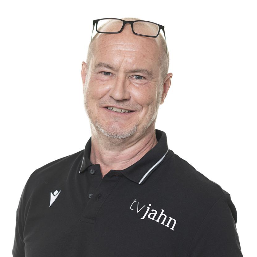 Porträt eines lächelnden Mannes mit Brille, der ein schwarzes Poloshirt mit dem Logo „tvjahn“ trägt.