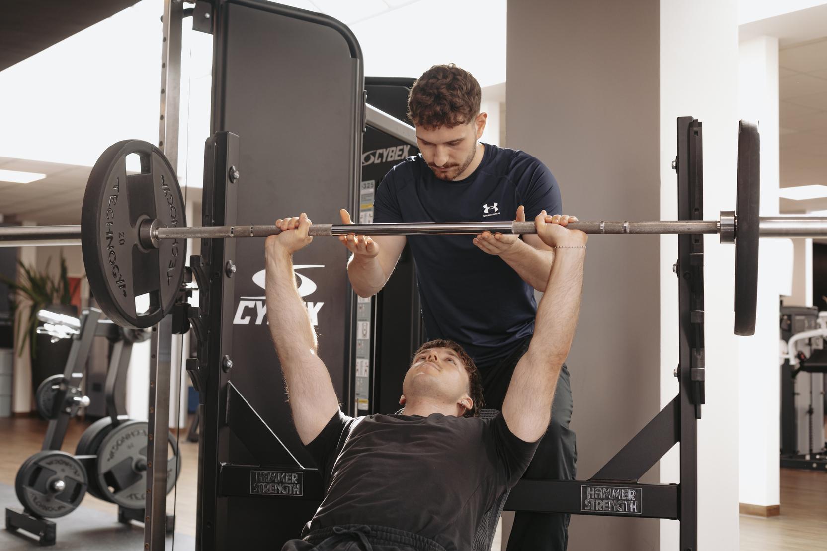 Trainingsszene im Fitnessstudio: Ein Trainer unterst&uuml;tzt einen Sportler beim Bankdr&uuml;cken mit einer Langhantel.