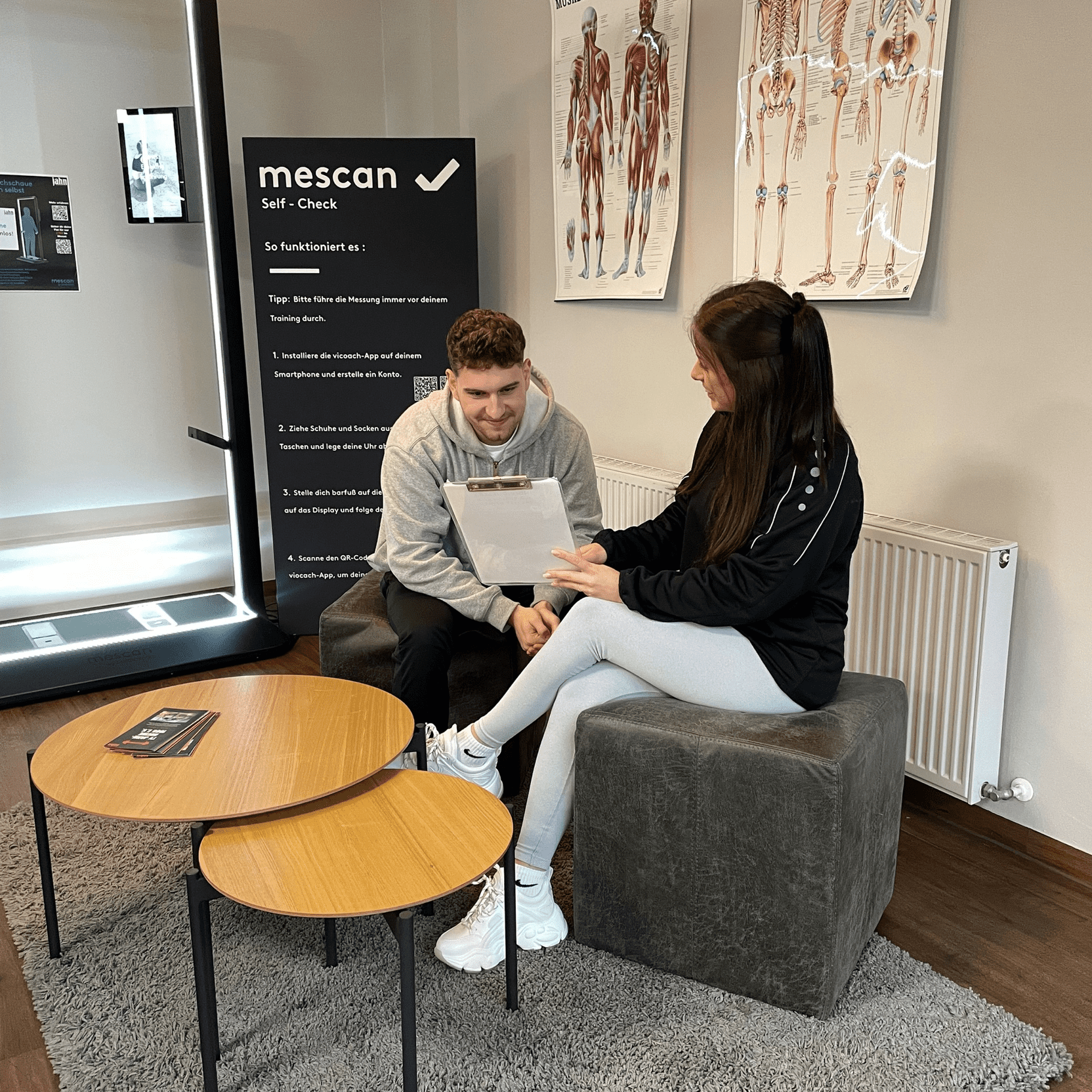 Zwei Personen sitzen in einem modernen Wartebereich und besprechen Unterlagen. Hintergrund mit anatomischen Postern.