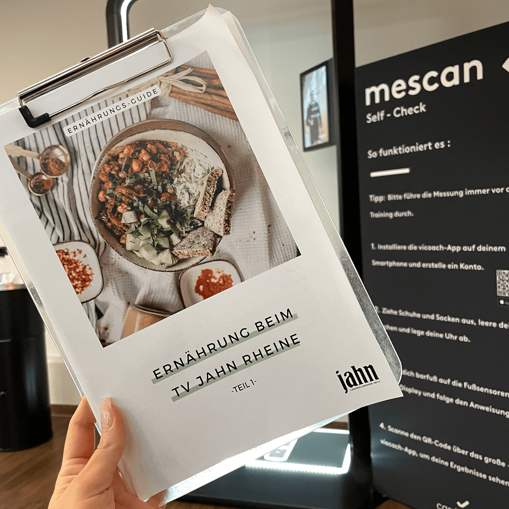 Hand hält einen Ernährungsleitfaden mit dem Titel „Ernährung beim TV Jahn Rheine“ vor einem Spiegel und QR-Code-Wand.