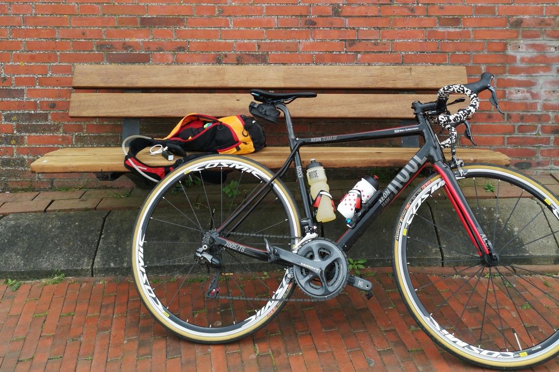 Schwarzes Rennrad mit buntem Rucksack und Zubeh&ouml;r auf einer Holzb&auml;nke vor einer roten Ziegelsteinmauer.