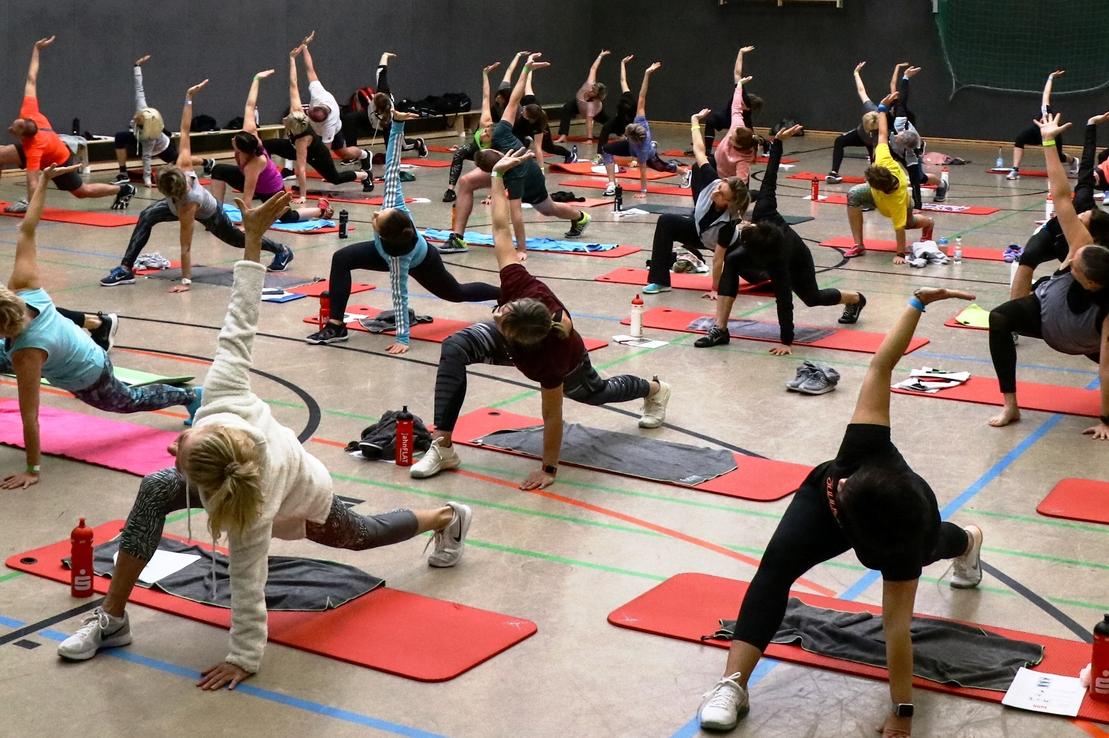 Gruppentraining mit vielen Teilnehmern auf Yogamatten in einer Halle, verschiedene Dehn- und Fitnesspositionen.
