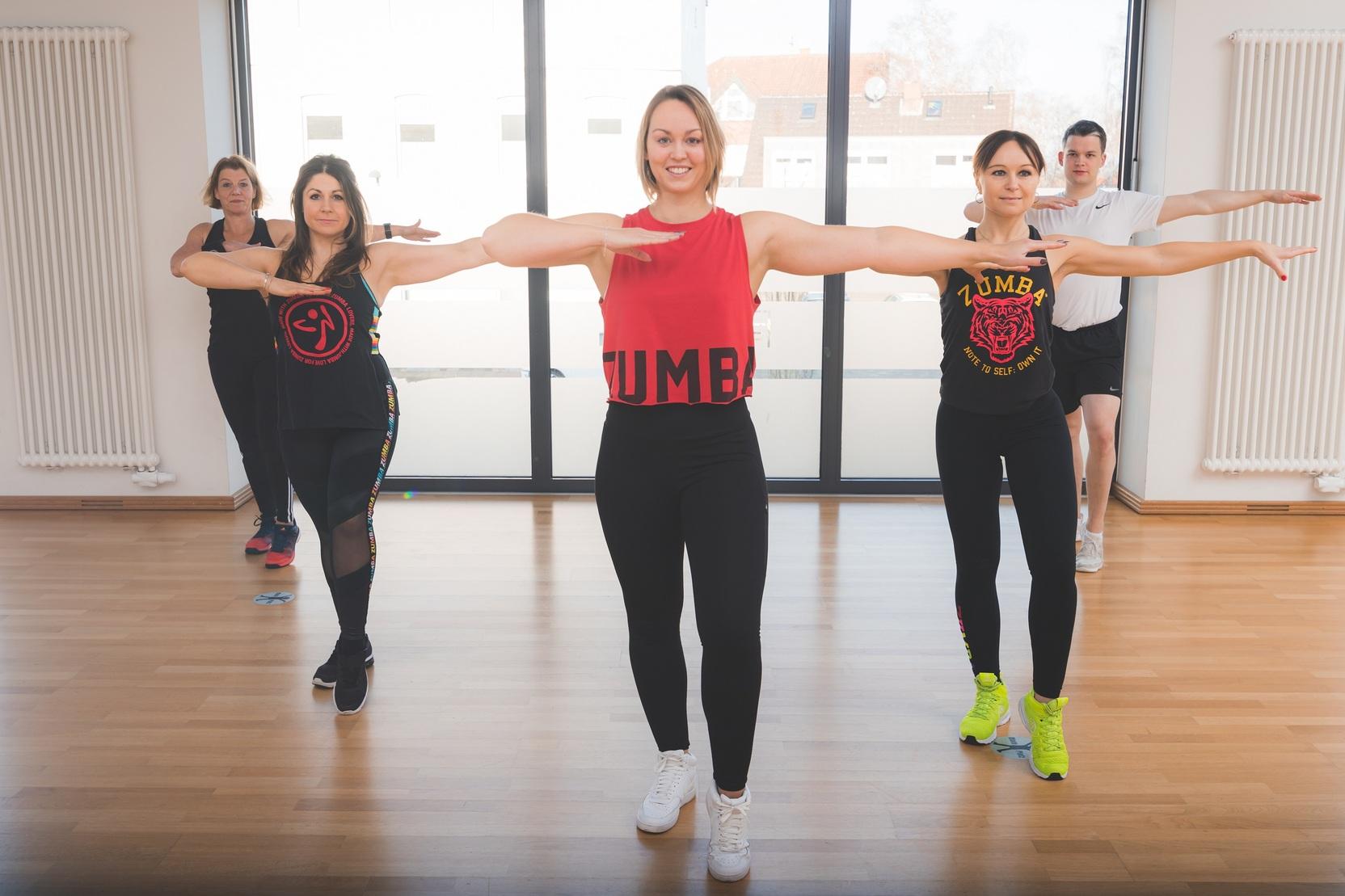 Gruppentraining mit f&uuml;nf Personen in sportlicher Kleidung, die synchron Bewegungen in einem hellen Raum ausf&uuml;hren.