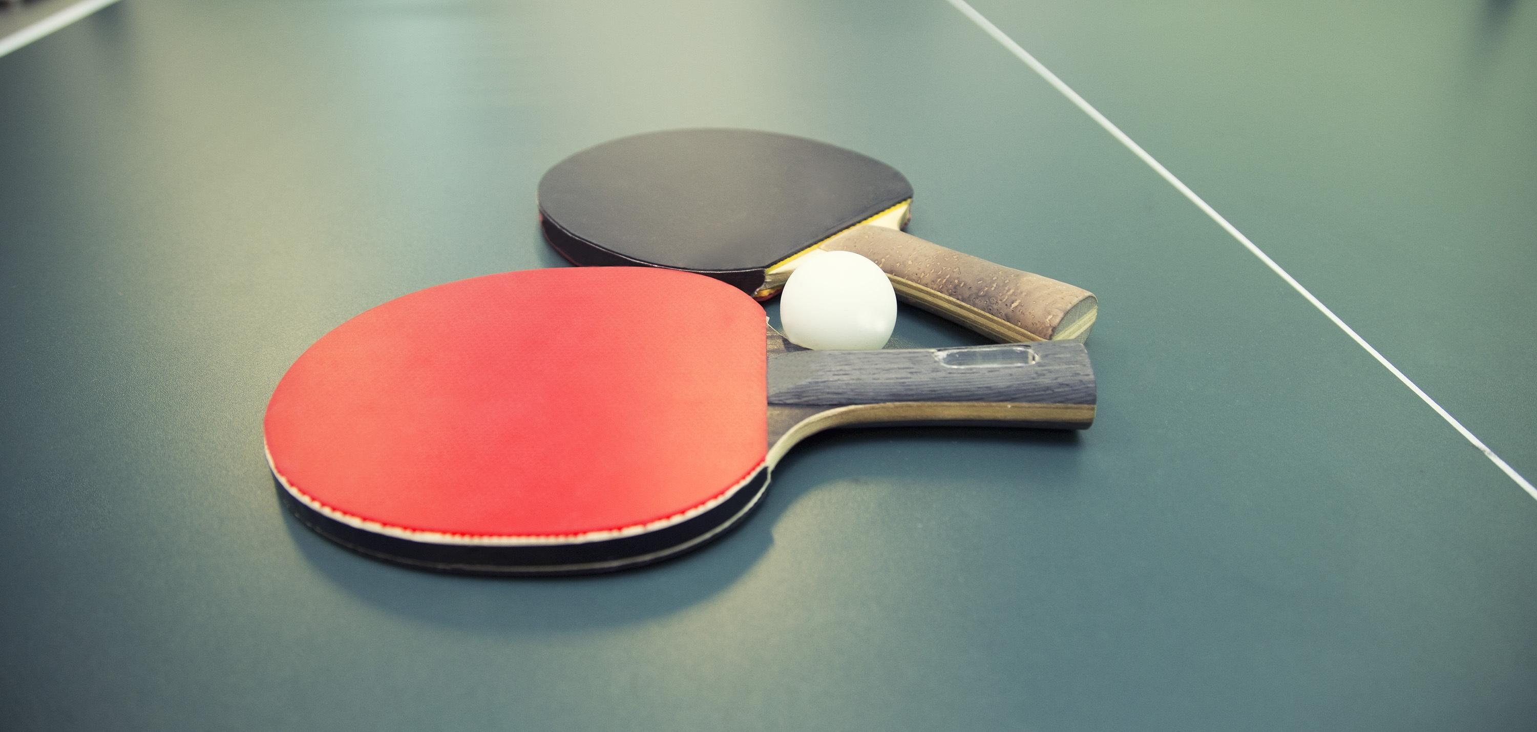 Tischtennisschl&auml;ger in Rot und Schwarz sowie ein Tischtennisball auf einer gr&uuml;nen Tischtennisplatte.
