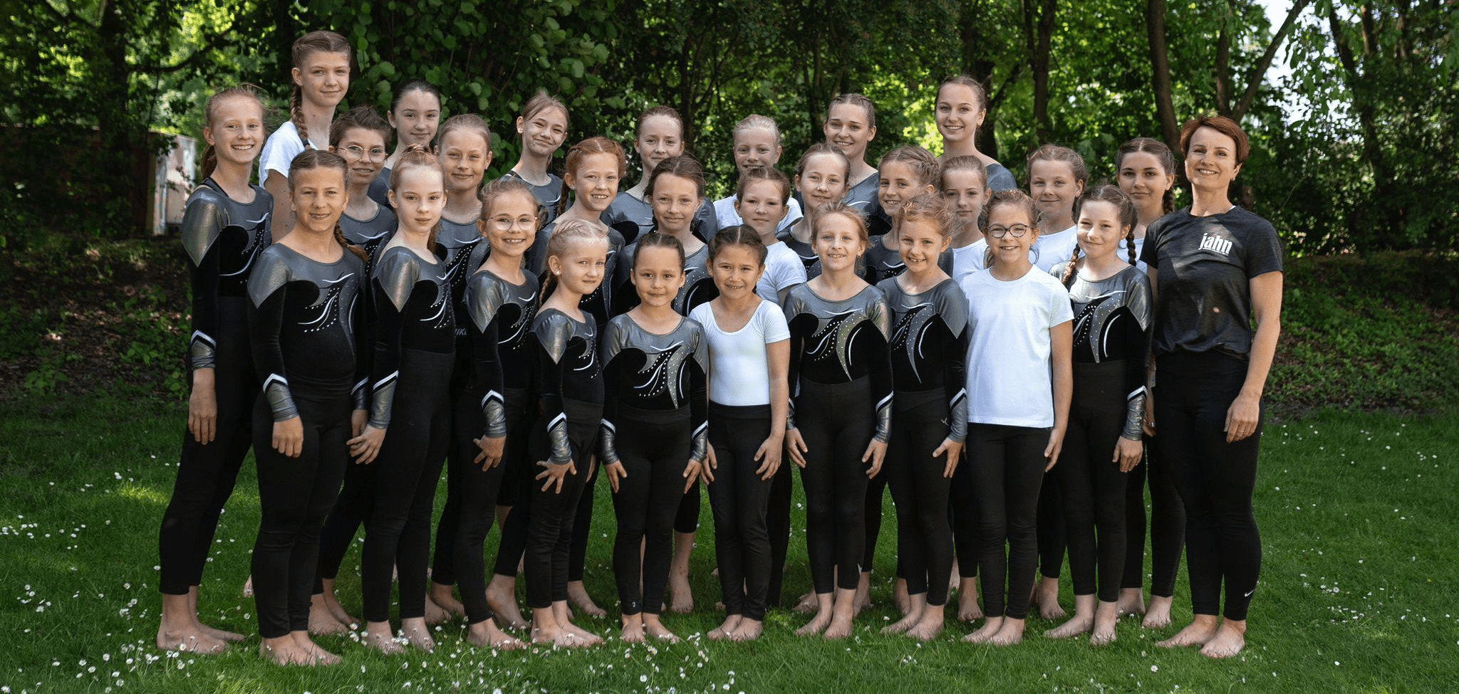 Eine Gruppe von 24 Gymnastinnen in schwarzen Outfits posiert in einem Garten, begleitet von einer Trainerin.
