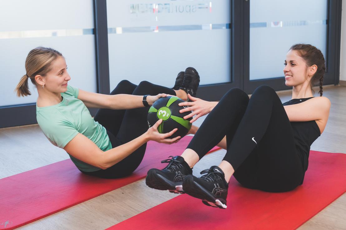 Zwei Frauen in Sportkleidung &uuml;ben auf roten Matten mit einem Medizinball in einem hellen Fitnessstudio.