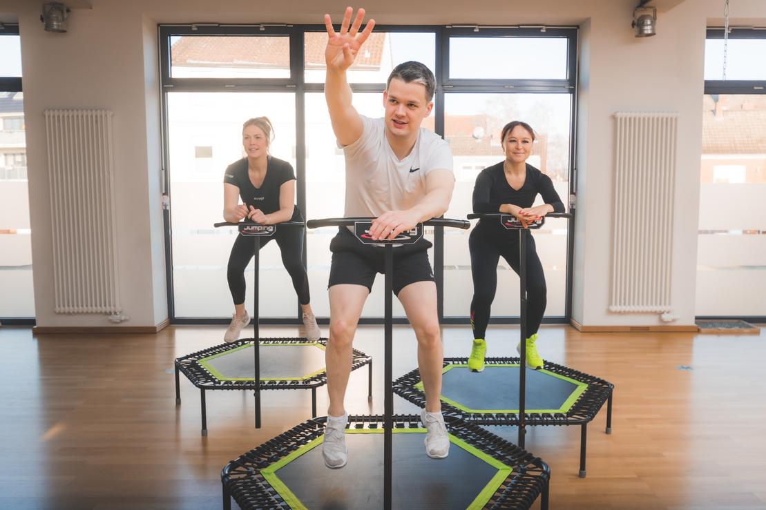Drei Personen beim Trampolinspringen in einem hellen Fitnessraum mit gro&szlig;en Fenstern.